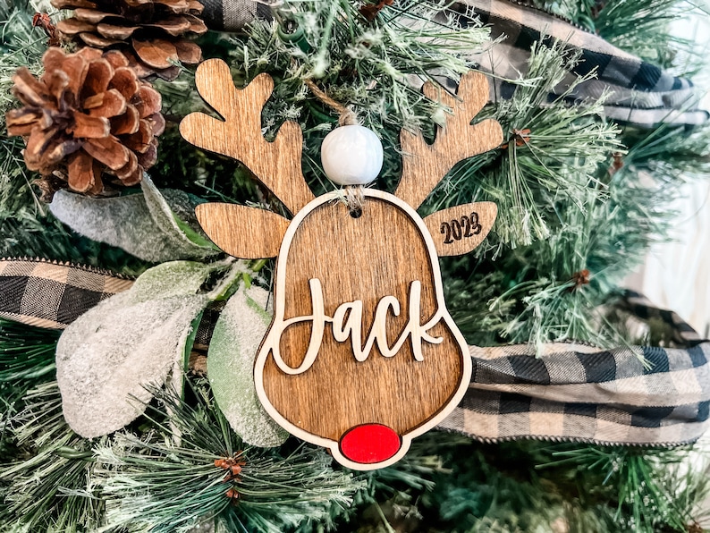 May include: Wooden reindeer ornament with a red nose, white bead, and the name "Jack" written on it. The ornament also includes the year "2023" on the side.