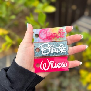 May include: Three personalized name tags in the image. The top tag is tie-dye with the name "Charlie" in pink. The middle tag is glittery blue with the name "Birdie" in white. The bottom tag is pink with the name "Willow" in white.