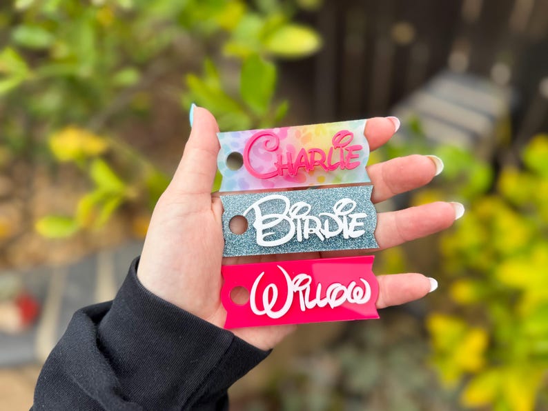 May include: Three personalized name tags in the image. The top tag is tie-dye with the name "Charlie" in pink. The middle tag is glittery blue with the name "Birdie" in white. The bottom tag is pink with the name "Willow" in white.