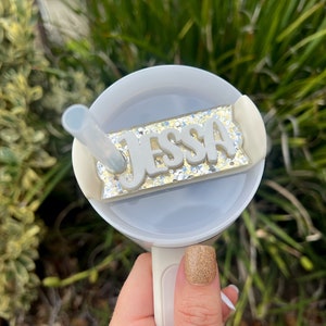 Op de afbeelding: Een witte beker met een doorzichtig deksel en een rietje. Het deksel heeft een gouden glitterachtergrond met een wit naamschild waarop "Jessa" staat.