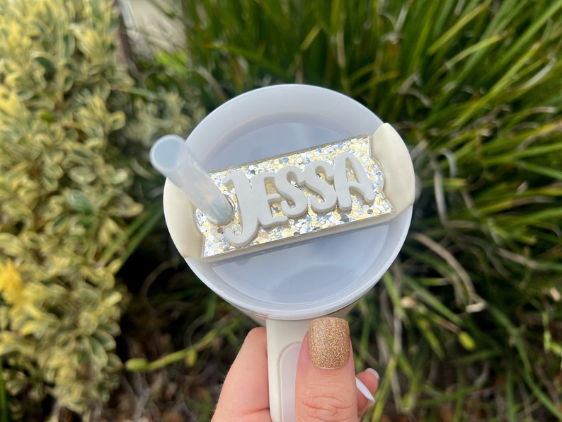 Op de afbeelding: Een witte beker met een doorzichtig deksel en een rietje. Het deksel heeft een gouden glitterachtergrond met een wit naamschild waarop "Jessa" staat.