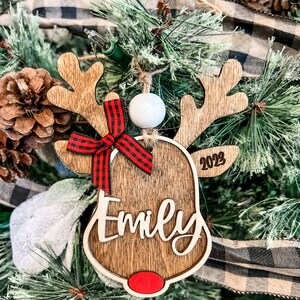 May include: Wooden reindeer Christmas ornament with a red and black plaid bow, a white bead, and the name "Emily" engraved on the front. The ornament also includes the year "2023" on the side.