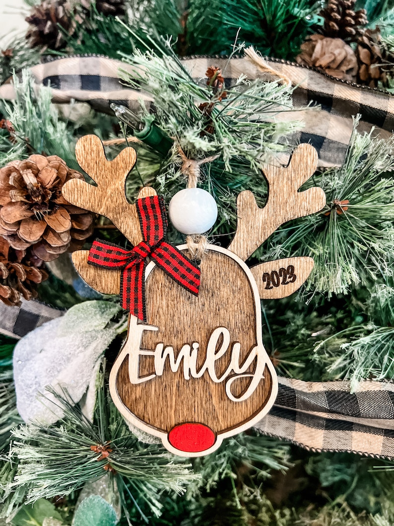 May include: Wooden reindeer Christmas ornament with a red and black plaid bow, a white bead, and the name "Emily" engraved on the front. The ornament also includes the year "2023" on the side.