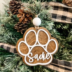 May include: Wooden paw print ornament with the name "Sadie" in white script. The ornament is brown with a white paw print outline and a white bead for hanging. It is displayed on a Christmas tree with pine needles, pine cones, and a black and white plaid ribbon.