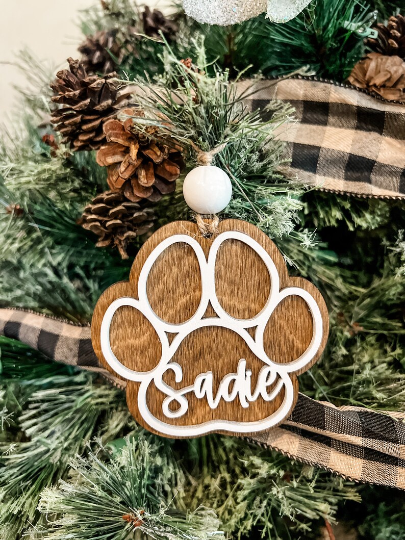 May include: Wooden paw print ornament with the name "Sadie" in white script. The ornament is brown with a white paw print outline and a white bead for hanging. It is displayed on a Christmas tree with pine needles, pine cones, and a black and white plaid ribbon.