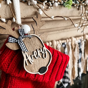Op de afbeelding: Een houten rendier ornament met een zwart-wit geblokte strik en de naam "Avery" erop geschreven. Het ornament hangt aan een rode gebreide sok.