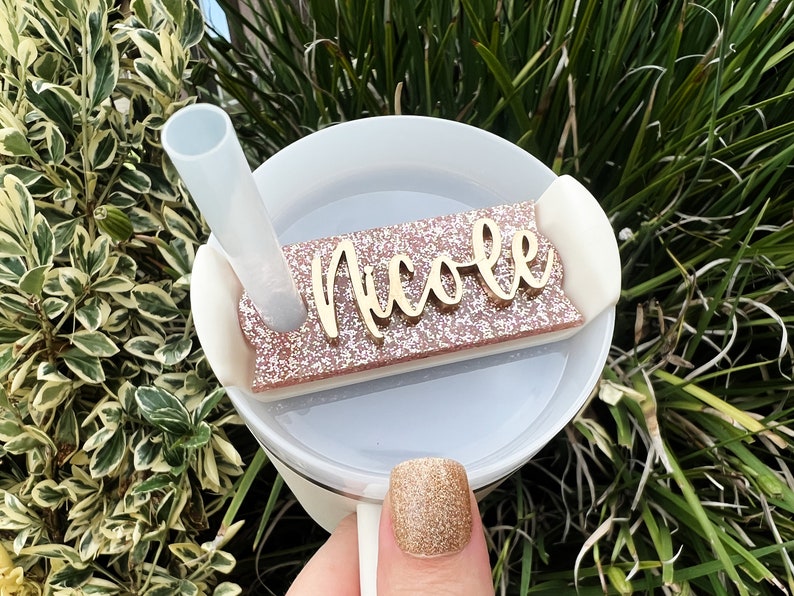 Op de afbeelding: Een witte tumbler met een roze glitterdeksel en een gouden naamplaatje met de tekst "Nicole".