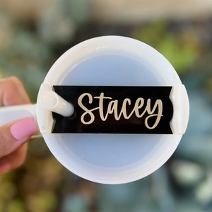 Op de afbeelding: Een witte tumbler met een zwart naamschild waarop "Stacey" in gouden letters staat. De tumbler heeft een doorzichtig deksel en een rietje.