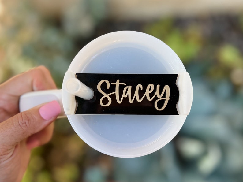 Op de afbeelding: Een witte tumbler met een zwart naamschild waarop "Stacey" in gouden letters staat. De tumbler heeft een doorzichtig deksel en een rietje.