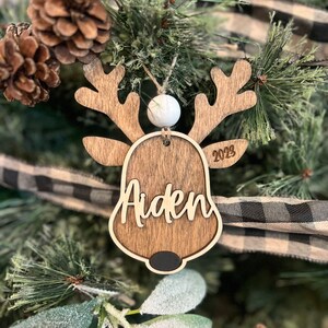 May include: Wooden reindeer ornament with the name Aiden and the year 2023 engraved on it. The ornament has a white bead at the top and a black nose.