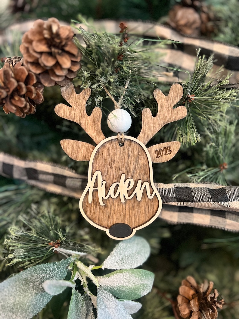 May include: Wooden reindeer ornament with the name Aiden and the year 2023 engraved on it. The ornament has a white bead at the top and a black nose.