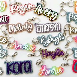 May include: A collection of colorful, glittery, and patterned keychains with various names written on them. The keychains are made of acrylic and have a variety of designs, including glitter, leopard print, and rainbow.