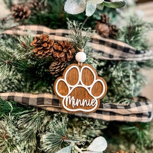 May include: A wooden Christmas ornament shaped like a paw print, with the name "Minnie" in white script. The ornament is hanging on a Christmas tree decorated with pinecones and a black and white plaid ribbon.