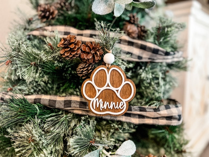 May include: A wooden Christmas ornament shaped like a paw print, with the name "Minnie" in white script. The ornament is hanging on a Christmas tree decorated with pinecones and a black and white plaid ribbon.