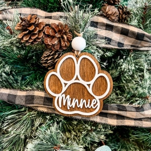 May include: A wooden paw print Christmas ornament with the name "Minnie" in white script. The ornament hangs from a white bead and is set against a backdrop of a Christmas tree with pinecones and a black and white plaid ribbon.