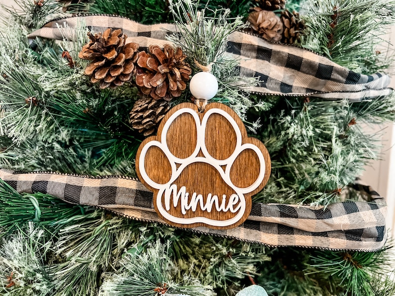 May include: A wooden paw print Christmas ornament with the name "Minnie" in white script. The ornament hangs from a white bead and is set against a backdrop of a Christmas tree with pinecones and a black and white plaid ribbon.