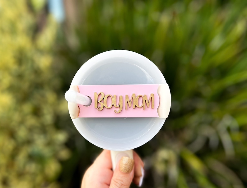 Op de afbeelding: Een witte beker met een roze en gouden "Boy Mom"-label bevestigd aan de deksel.