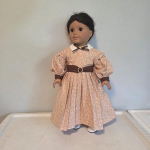 May include: A doll wearing a peach-colored dress with a floral pattern. The dress has a white collar, brown belt with a gold buckle, and puffed sleeves. The doll has dark hair and is wearing small gold earrings.