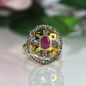 May include: A gold ring with a central oval ruby, surrounded by a halo of small diamonds and green and yellow gemstones. The ring has a floral design and is set against a blurred background.
