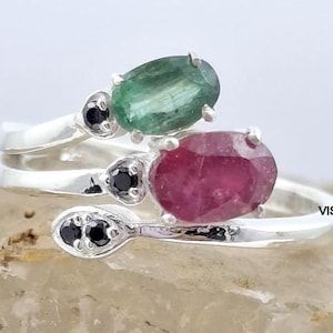May include: A silver ring with a bypass design, featuring a green oval gemstone and a red oval gemstone. The ring also has small black gemstones. The ring is on a light brown surface.