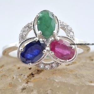 Pode incluir: Um anel de prata com três pedras preciosas ovais: uma esmeralda verde, uma safira azul e um rubi rosa. As pedras são engastadas num design floral, com pequenas pedras transparentes. O anel é exibido numa superfície castanha clara.