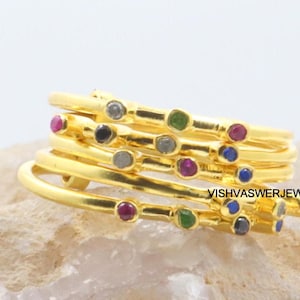 Op de afbeelding: Een stapel goudkleurige ringen, elk versierd met kleine, kleurrijke edelstenen. De edelstenen zijn in verschillende kleuren, waaronder rood, groen, blauw en grijs. De ringen zijn op elkaar gestapeld, waardoor een gelaagd effect ontstaat. De tekst "VISHVASWERJEWELLERS" is zichtbaar.