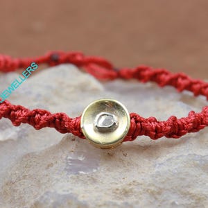 May include: A red string bracelet with a gold round charm featuring a clear diamond. The bracelet is on a light gray rock.