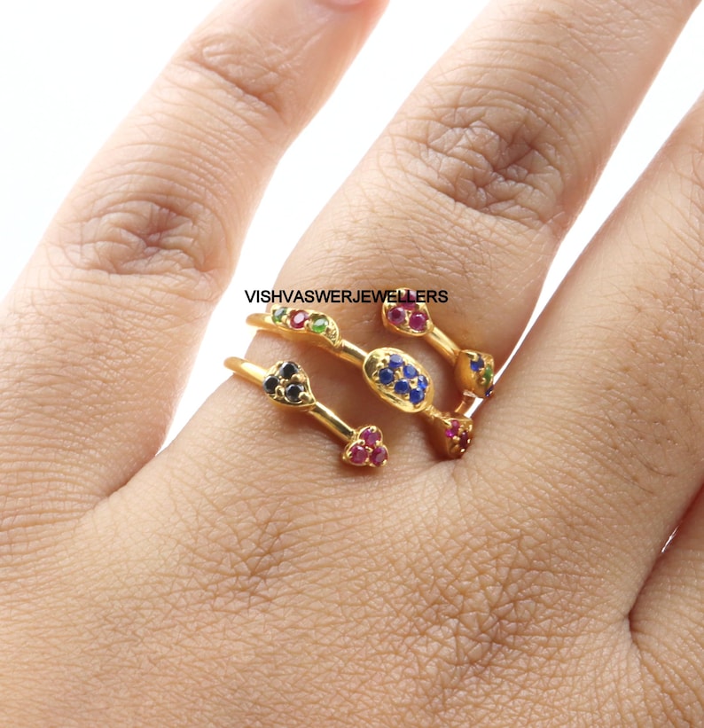 Pu&ograve; includere: Un anello dorato con pi&ugrave; fasce, ognuna ornata di pietre preziose colorate. L'anello presenta pietre rosse, blu, verdi e nere, creando un design vibrante. Il testo "VISHVASWERJEWELLERS" &egrave; visibile.