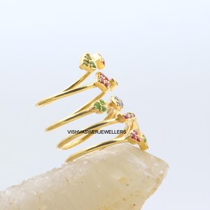 May include: A gold-colored spiral ring with small, colorful gemstones. The ring features a unique design with multiple loops, each adorned with clusters of red, green, and purple gemstones. The ring is set against a light background.