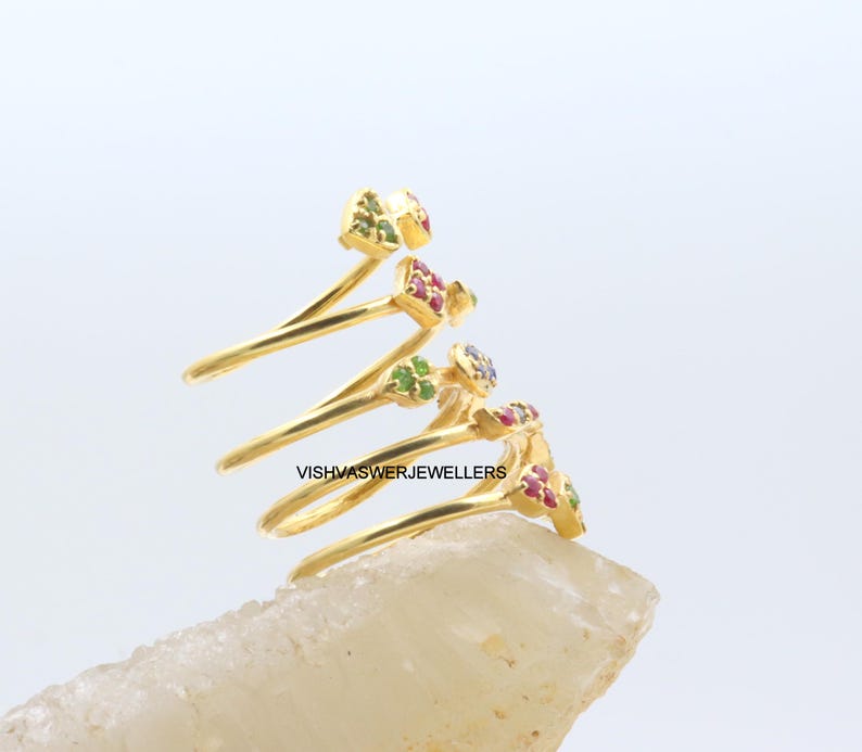 May include: A gold-colored spiral ring with small, colorful gemstones. The ring features a unique design with multiple loops, each adorned with clusters of red, green, and purple gemstones. The ring is set against a light background.