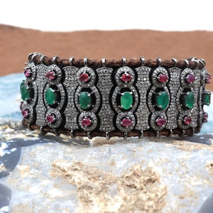 Puede incluir: Un brazalete de plata con una banda de cuero marrón, con piedras preciosas de color verde esmeralda y rojo rubí engastadas en un diseño de pavé de diamantes.