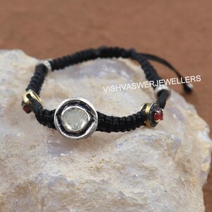 Peut inclure: Un bracelet tressé noir avec un pendentif en pierre précieuse argent et blanc et des accents de pierres précieuses rouges. Le bracelet a une fermeture à nœud coulissant et le texte "VISHVASWERJEWELLERS" est visible sur le bracelet.