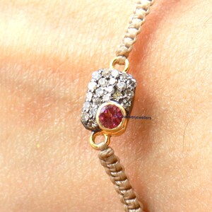 May include: A beige braided bracelet with a gold rectangular charm set with small white diamonds and a pink gemstone in the center.