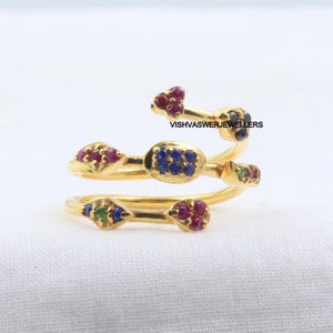 Pu&ograve; includere: Un anello color oro con un design a spirale, adornato con piccole pietre preziose colorate. L'anello presenta pietre rosse, blu e verdi e il testo "VISHVASWERJEWELLERS". L'anello &egrave; posto su uno sfondo bianco.