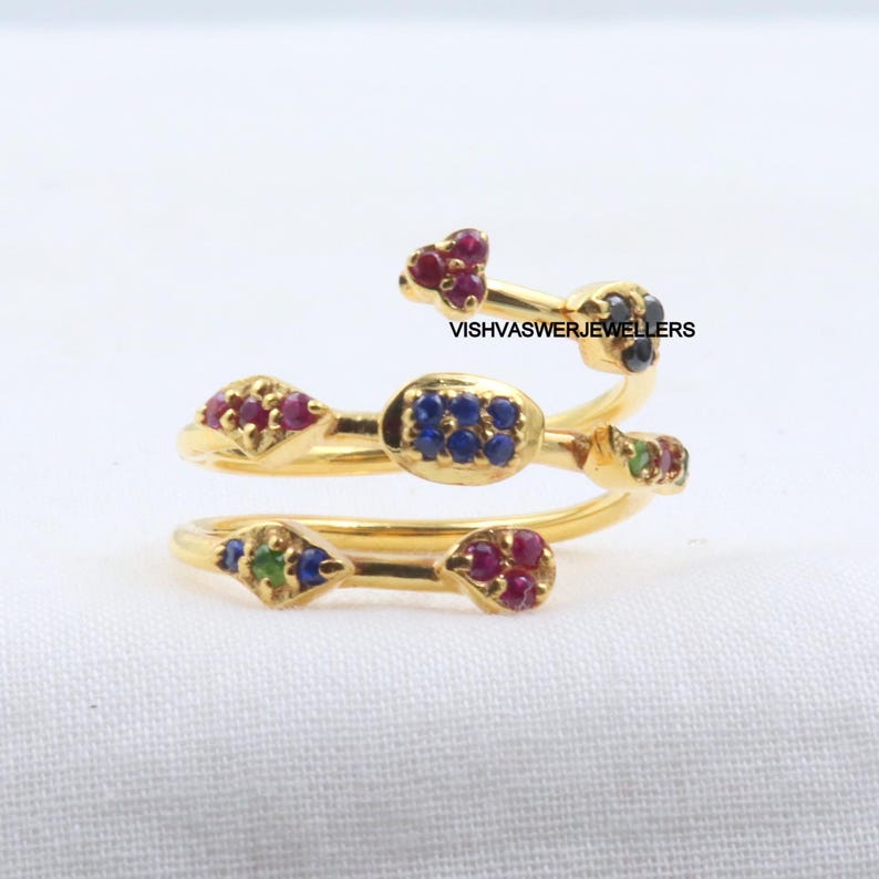 Pu&ograve; includere: Un anello color oro con un design a spirale, adornato con piccole pietre preziose colorate. L'anello presenta pietre rosse, blu e verdi e il testo "VISHVASWERJEWELLERS". L'anello &egrave; posto su uno sfondo bianco.