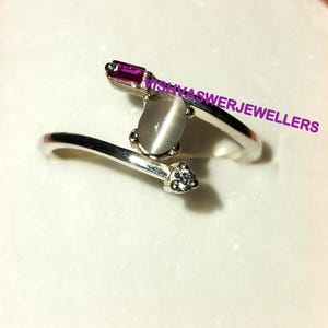 May include: A silver ring with a white oval gemstone, a small clear gemstone, and a pink rectangular gemstone. The ring is set on a white background.