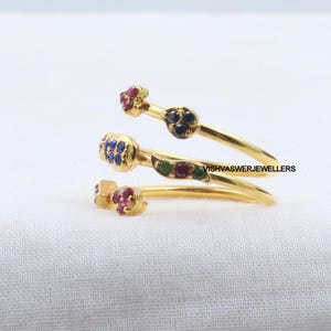 Pu&ograve; includere: Un anello avvolgente color oro con tre fasce a spirale. Ogni fascia presenta piccole pietre preziose colorate in rosso, blu, verde e nero. L'anello &egrave; posto su uno sfondo bianco.