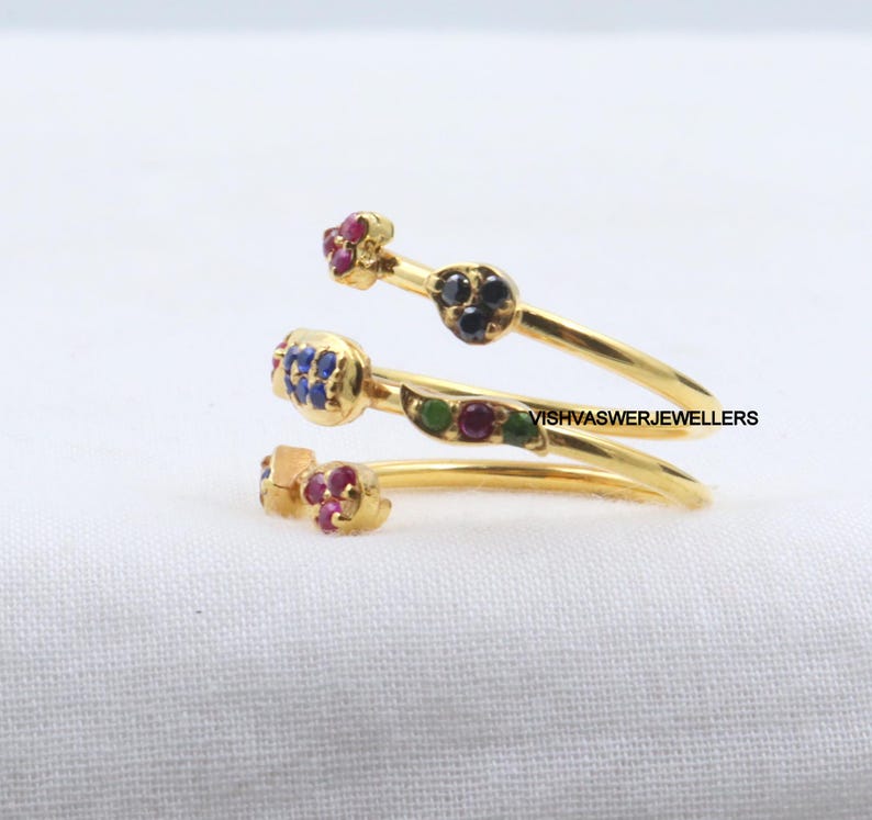 Pu&ograve; includere: Un anello avvolgente color oro con tre fasce a spirale. Ogni fascia presenta piccole pietre preziose colorate in rosso, blu, verde e nero. L'anello &egrave; posto su uno sfondo bianco.