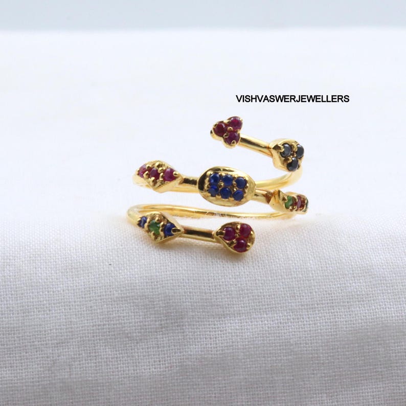 Pu&ograve; includere: Un anello a spirale color oro con accenti di pietre preziose colorate. L'anello presenta pietre preziose rosse, blu, verdi e nere. Il testo "VISHVASWERJEWELLERS" &egrave; visibile sullo sfondo.