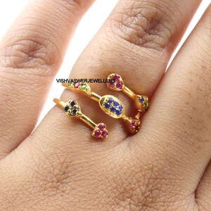 Pu&ograve; includere: Anello dorato con pi&ugrave; sezioni, ognuna ornata di pietre preziose colorate. L'anello presenta pietre rosse, blu, verdi e nere, creando un design vibrante. Il testo "VISHVASWERJEWELLERS" &egrave; visibile.