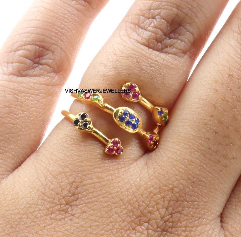 Pu&ograve; includere: Anello dorato con pi&ugrave; sezioni, ognuna ornata di pietre preziose colorate. L'anello presenta pietre rosse, blu, verdi e nere, creando un design vibrante. Il testo "VISHVASWERJEWELLERS" &egrave; visibile.