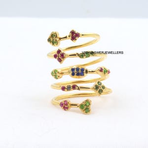 May include: A collection of gold-toned spiral rings, each adorned with colorful gemstones. The rings feature various shapes, including hearts, diamonds, and ovals, with green, red, and blue stones. The text "VISHVASWERJEWELLERS" is visible.