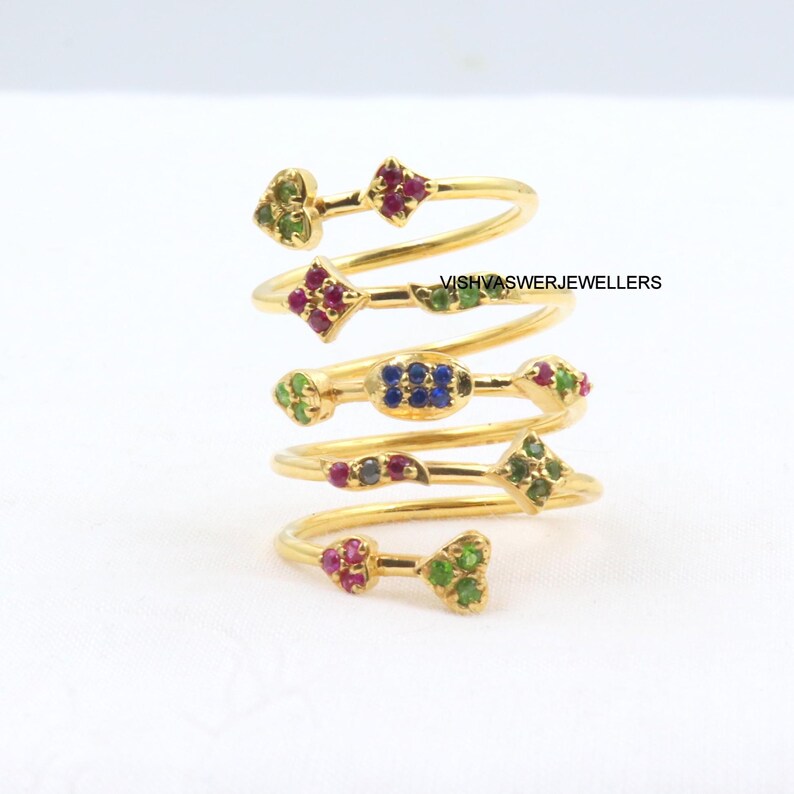 May include: A collection of gold-toned spiral rings, each adorned with colorful gemstones. The rings feature various shapes, including hearts, diamonds, and ovals, with green, red, and blue stones. The text "VISHVASWERJEWELLERS" is visible.