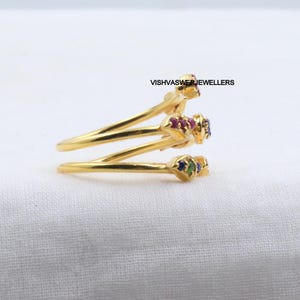 Pu&ograve; includere: Un anello color oro con un design a spirale. L'anello presenta tre sezioni, ciascuna ornata da piccole pietre preziose colorate. Le pietre preziose includono pietre rosse, blu e verdi. Il testo "VISHVASWERJEWELLERS" &egrave; visibile.