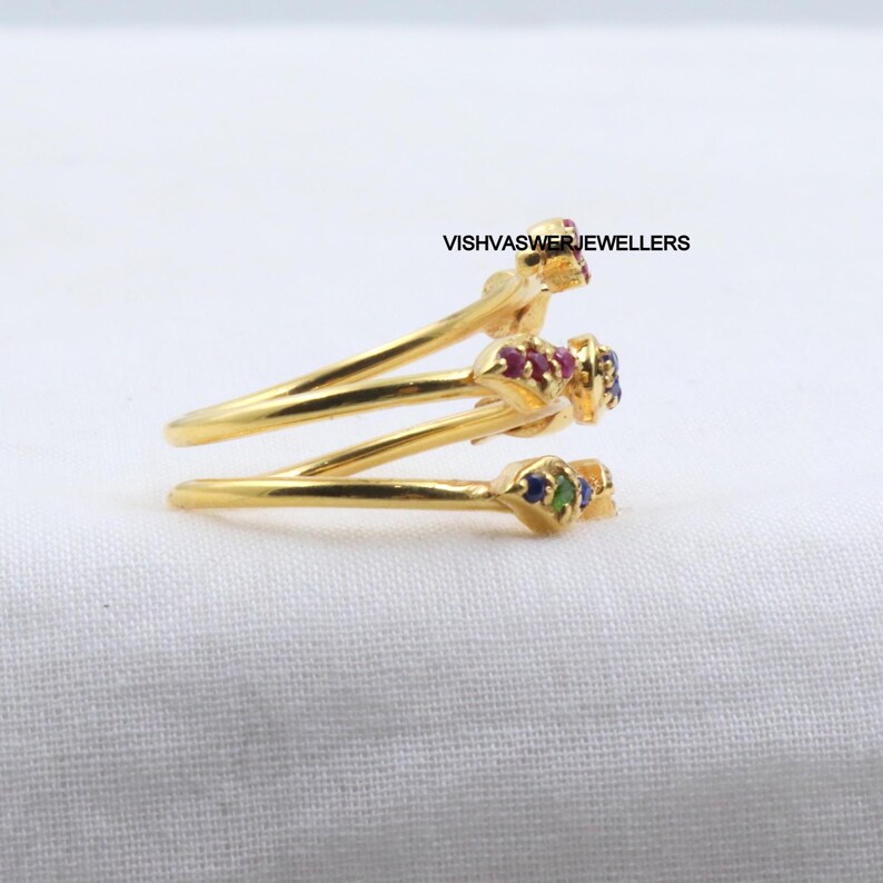 Pu&ograve; includere: Un anello color oro con un design a spirale. L'anello presenta tre sezioni, ciascuna ornata da piccole pietre preziose colorate. Le pietre preziose includono pietre rosse, blu e verdi. Il testo "VISHVASWERJEWELLERS" &egrave; visibile.