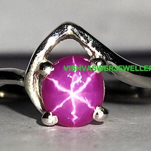 May include: A silver ring featuring a pink star ruby gemstone. The ring has a curved band and a prong setting for the stone.  "VISHVASVER JEWELLERS" is printed on the band.