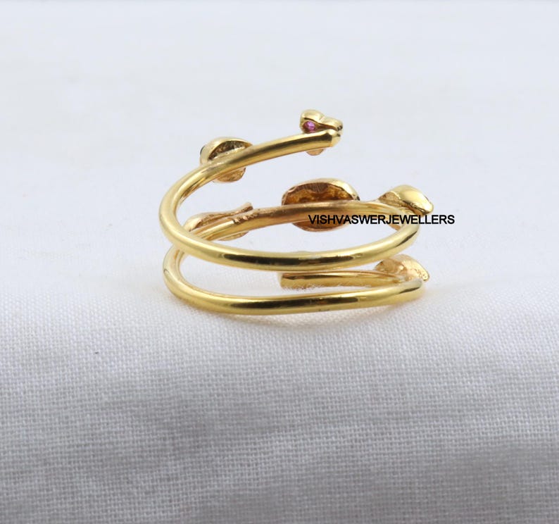 Pu&ograve; includere: Un anello color oro con un design a spirale. L'anello presenta piccoli dettagli a forma di foglia e minuscole pietre preziose rosa. Il testo "VISHVASWERJEWELLERS" &egrave; visibile sull'anello.