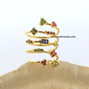 May include: A spiral-shaped gold ring with multiple bands, each adorned with colorful gemstones. The ring features green, red, and blue stones in various geometric shapes. The text "VISHVASWERJEWELLERS" is visible on the ring.