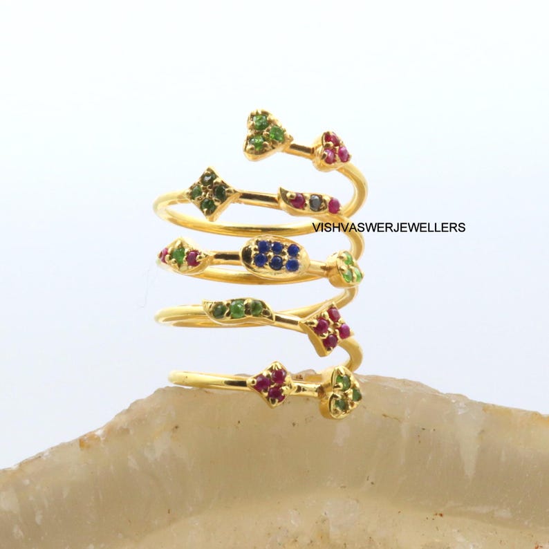 May include: A spiral-shaped gold ring with multiple bands, each adorned with colorful gemstones. The ring features green, red, and blue stones in various geometric shapes. The text "VISHVASWERJEWELLERS" is visible on the ring.