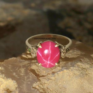 May include: A silver ring featuring a large, oval, pink gemstone with a star-like pattern. The ring has small accent stones on either side of the main stone. The text "VISHVASWERJEWELLERS" is visible on the ring.